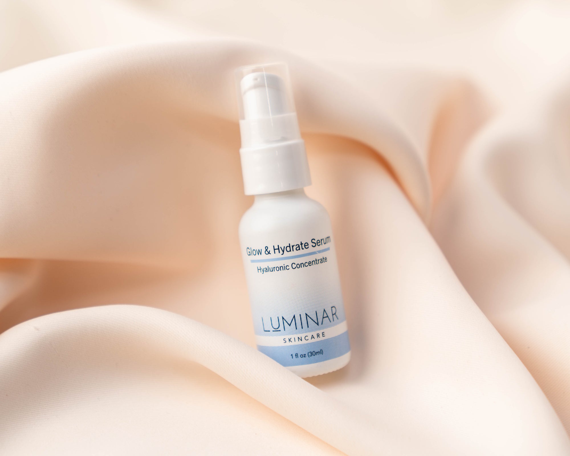 Luminar Glow & Hydrate Hyaluronic Serum bottle lying on soft peach fabric, highlighting deep hydration and radiance.